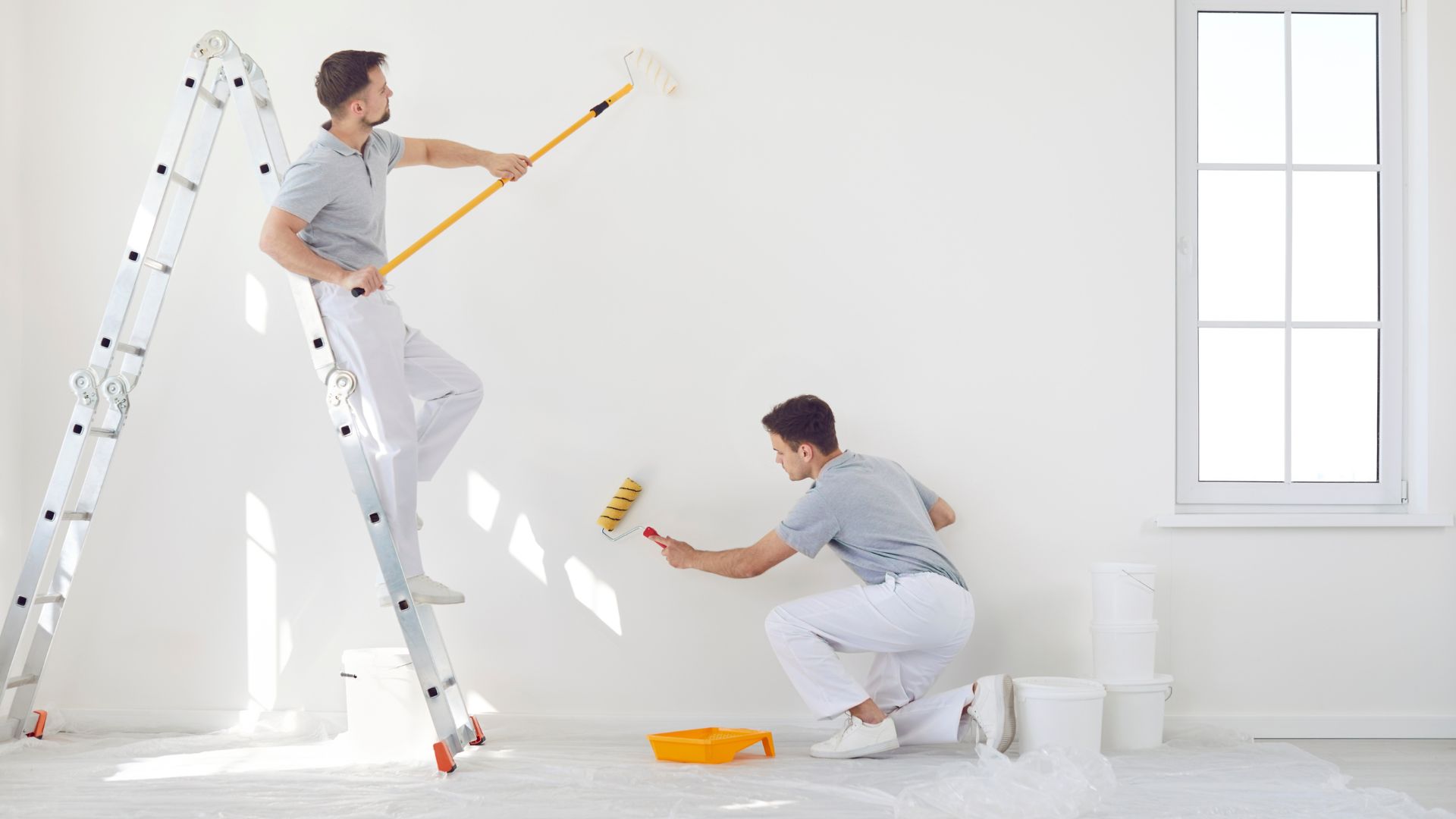 Two professional painters in white overalls working indoors: one on ladder rolling ceiling, one crouching to paint lower wall. Bright, clean prep.